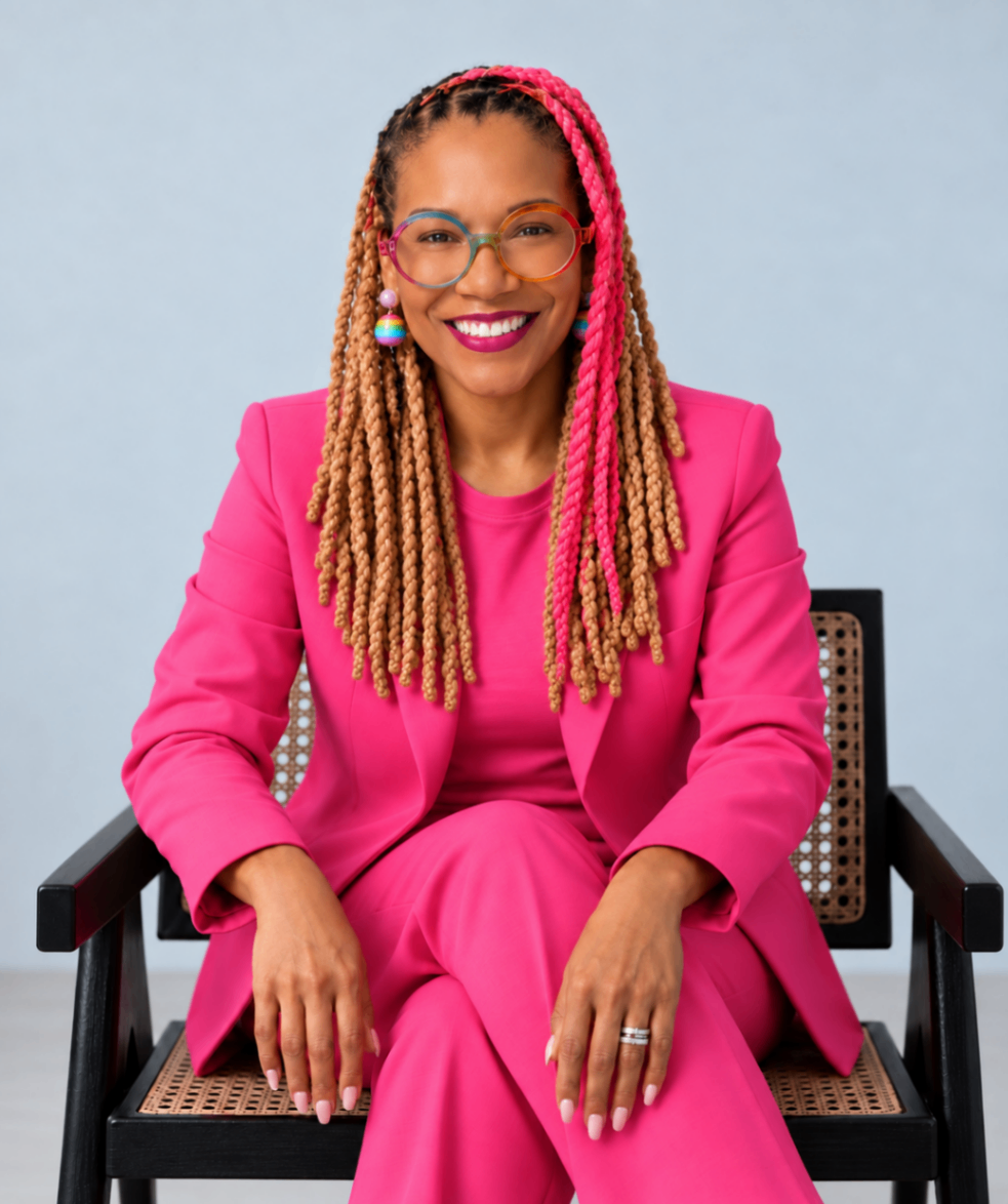 Angela Briggs-Paige seated from head to knees in a black-and-cane chair, wearing a hot-pink suit against a pale grey-blue background